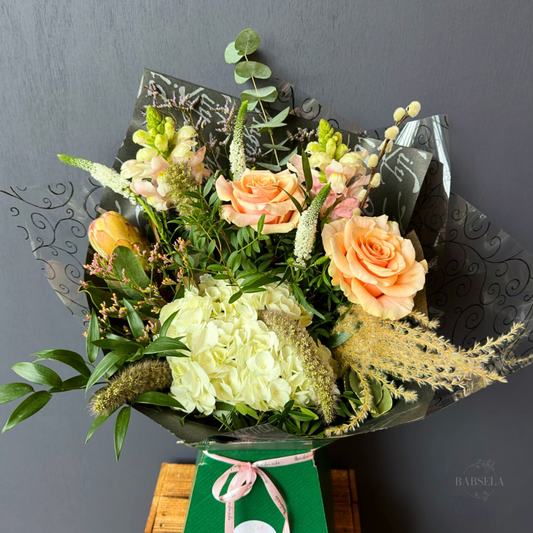 Elegant bouquet featuring peach roses, white hydrangeas, snapdragons, and greenery, wrapped in decorative black paper with a green box tied with a pink ribbon