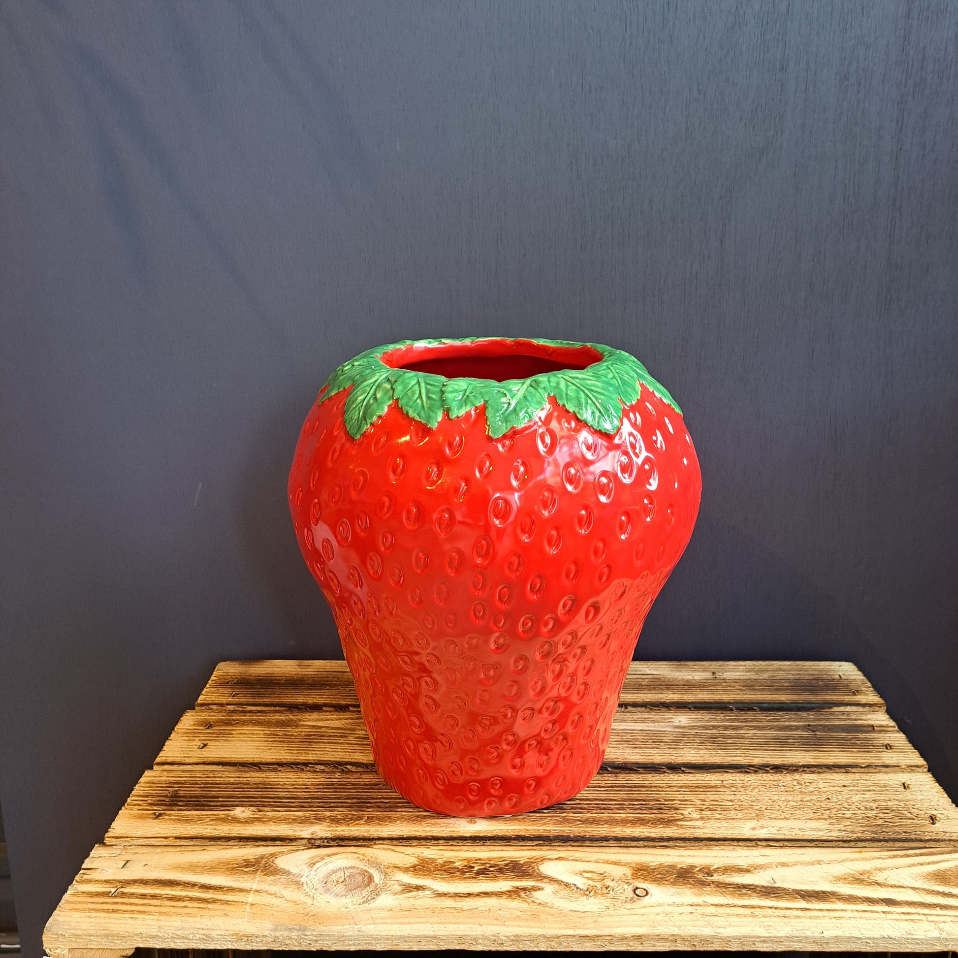 Ceramic vase shaped and painted like a large red strawberry with green leaves around the rim, placed on a wooden surface