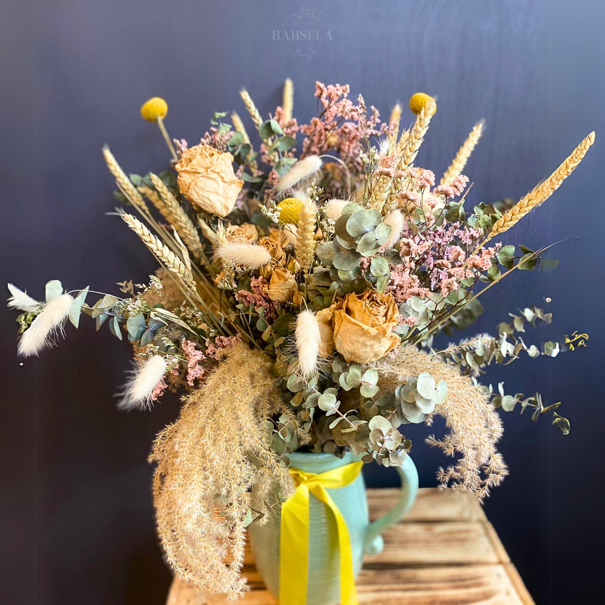 A rustic bouquet featuring dried flowers, wheat stalks, pampas grass, and eucalyptus leaves, arranged in a green pitcher tied with a yellow ribbon, placed on a wooden surface