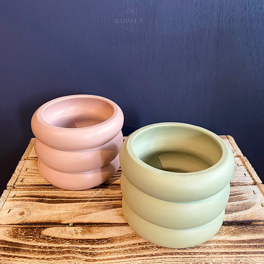 Two stylish, round ceramic plant pots with a ribbed texture, one in a soft pink color and the other in a pastel green shade, displayed on a rustic wooden surface against a dark blue background