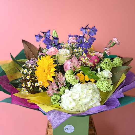 Vibrant mixed flower bouquet with sunflowers, roses, and delphiniums in colorful wrapping from Babsela Flowers