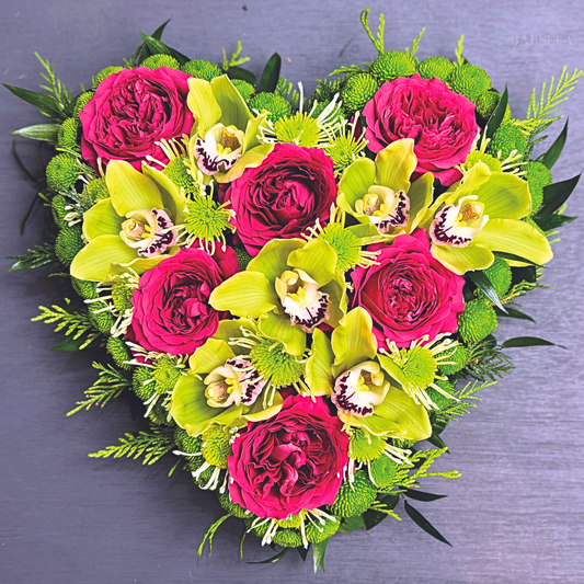 A heart-shaped floral arrangement featuring vibrant pink roses and green orchids, complemented by lush green foliage and small green accents, set against a dark background