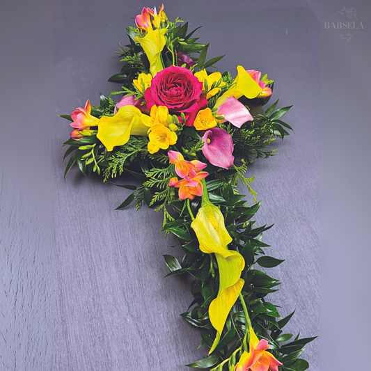 Floral cross arrangement featuring vibrant yellow calla lilies, pink roses, and mixed greenery, designed by Babsela