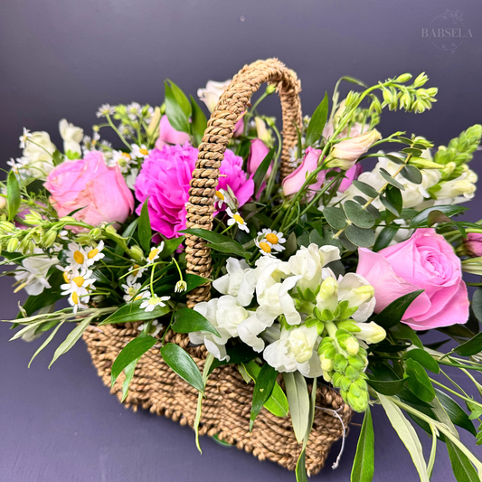 Woven basket filled with a vibrant arrangement of pink roses, white snapdragons, daisies, and green foliage, displayed against a dark background