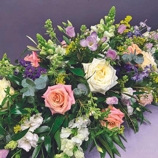 A vibrant floral arrangement featuring a mix of roses in pastel colors, white snapdragons, purple lisianthus, yellow solidago, and lush green foliage, set against a purple background