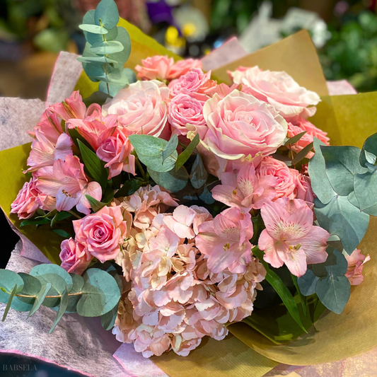 A bouquet featuring a mix of soft pink roses, alstroemeria, and hydrangeas, accented with green eucalyptus leaves, wrapped in pastel-colored paper