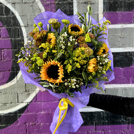 A bouquet of sunflowers and yellow wildflowers, accented with greenery and wrapped in purple paper with a yellow ribbon, held against a vibrant graffiti wall background