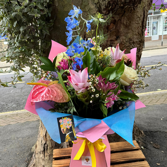 A colorful bouquet of mixed flowers, including blue delphiniums, pink lilies, white roses, and anthuriums, wrapped in pink and blue paper with a yellow ribbon, displayed outdoors near a tree