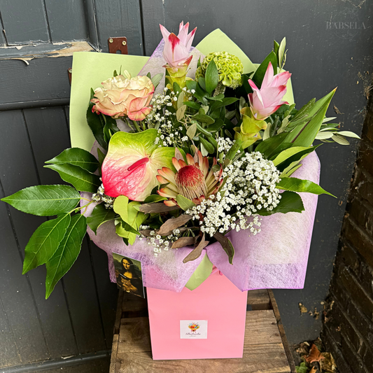 A lush bouquet of flowers arranged in a pink gift box, featuring pink curcuma flowers, green hydrangea, pink anthurium, protea, baby's breath, and assorted greenery, set against a rustic dark gray wooden background