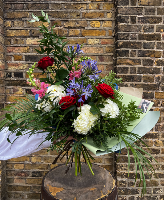 Elegant hand-tied bouquet with red roses, white hydrangeas, purple agapanthus, and greenery against a brick wall backdrop from Babsela Flowers